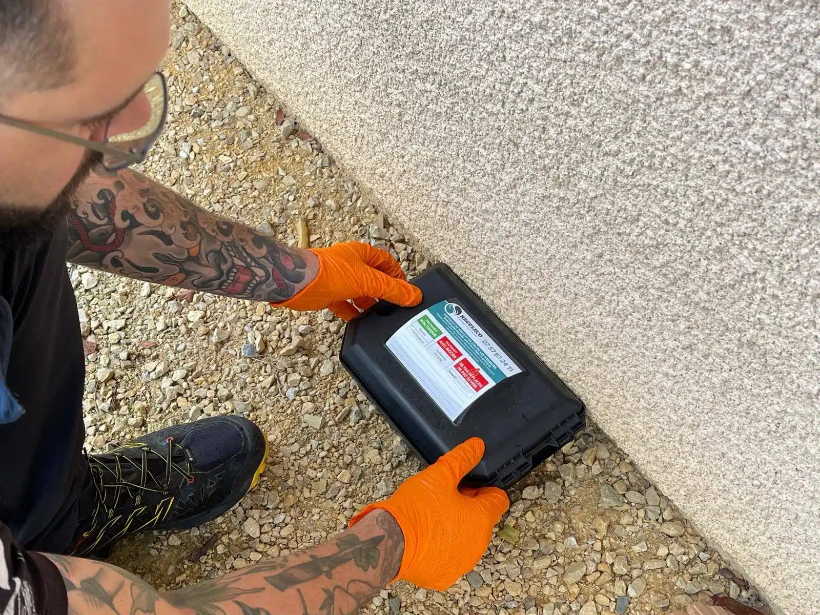 Technicien tatoué en gants orange plaçant une boîte de dératisation Risqueleco contre un mur. Raticide, Tél. 07 57 67 24 11 visible.